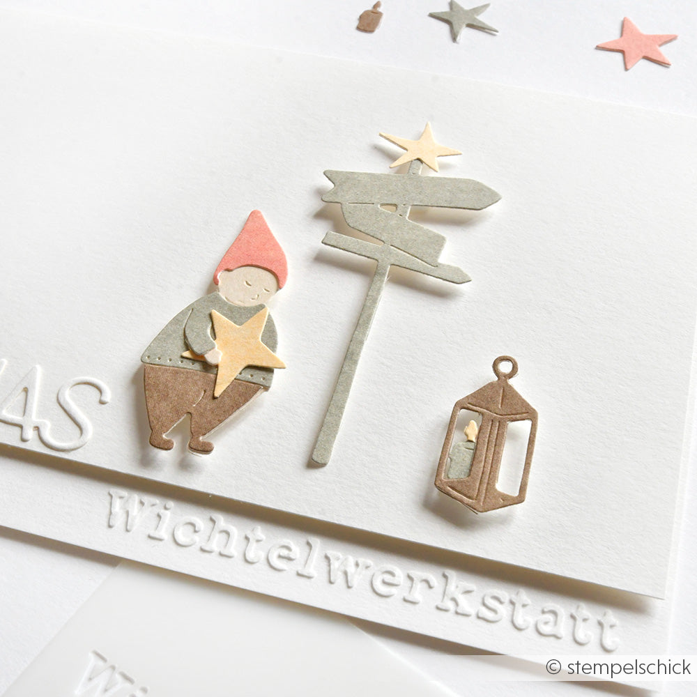 Scherenschnitte eines Gnoms, eines Wegweisers, einer Laterne und von Sternen auf einer weißen Strukturkarte mit der Aufschrift Wichtelwerkstatt von Alexandra Renke-perfekt als wiederverwendbare Schablonen für kreative Bastelideen. Stencil Magie für jedes Werk!.