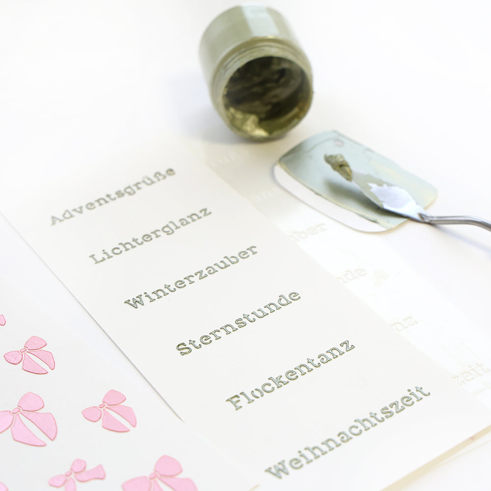 Ein Spachtel von Alexandra Renke, ein grüner Farbtopf und wiederverwendbare Schablonen mit deutschen Feiertagswörtern, die auf weißes Papier schabloniert sind.
