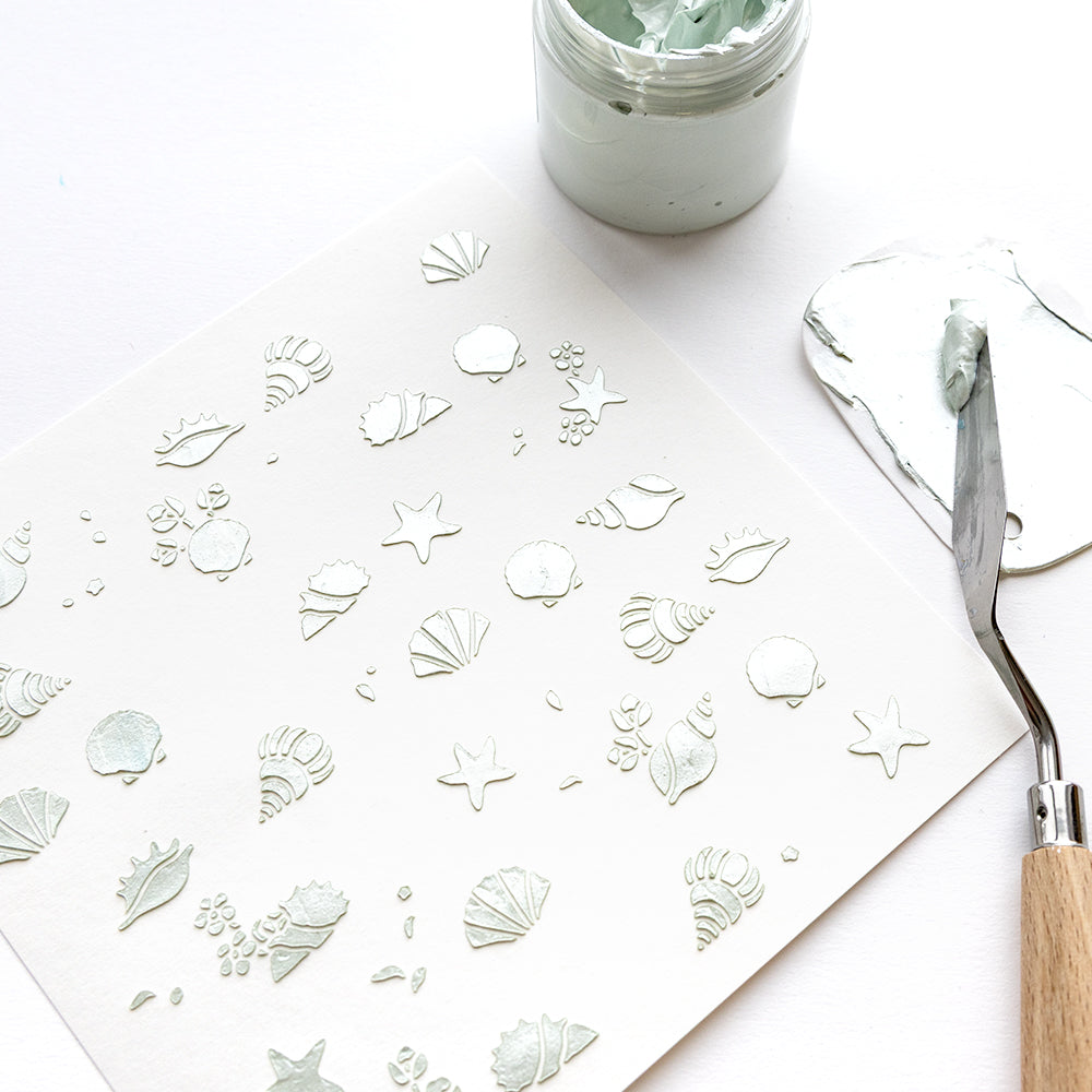 Blatt mit strukturierten Muschel- und Seesternmustern, hergestellt mit der Alexandra Renke Schablone Schablonen Florale Muscheln, ausgestellt neben einem Glas mintgrüner Paste und einem Spachtel auf weißem Hintergrund.