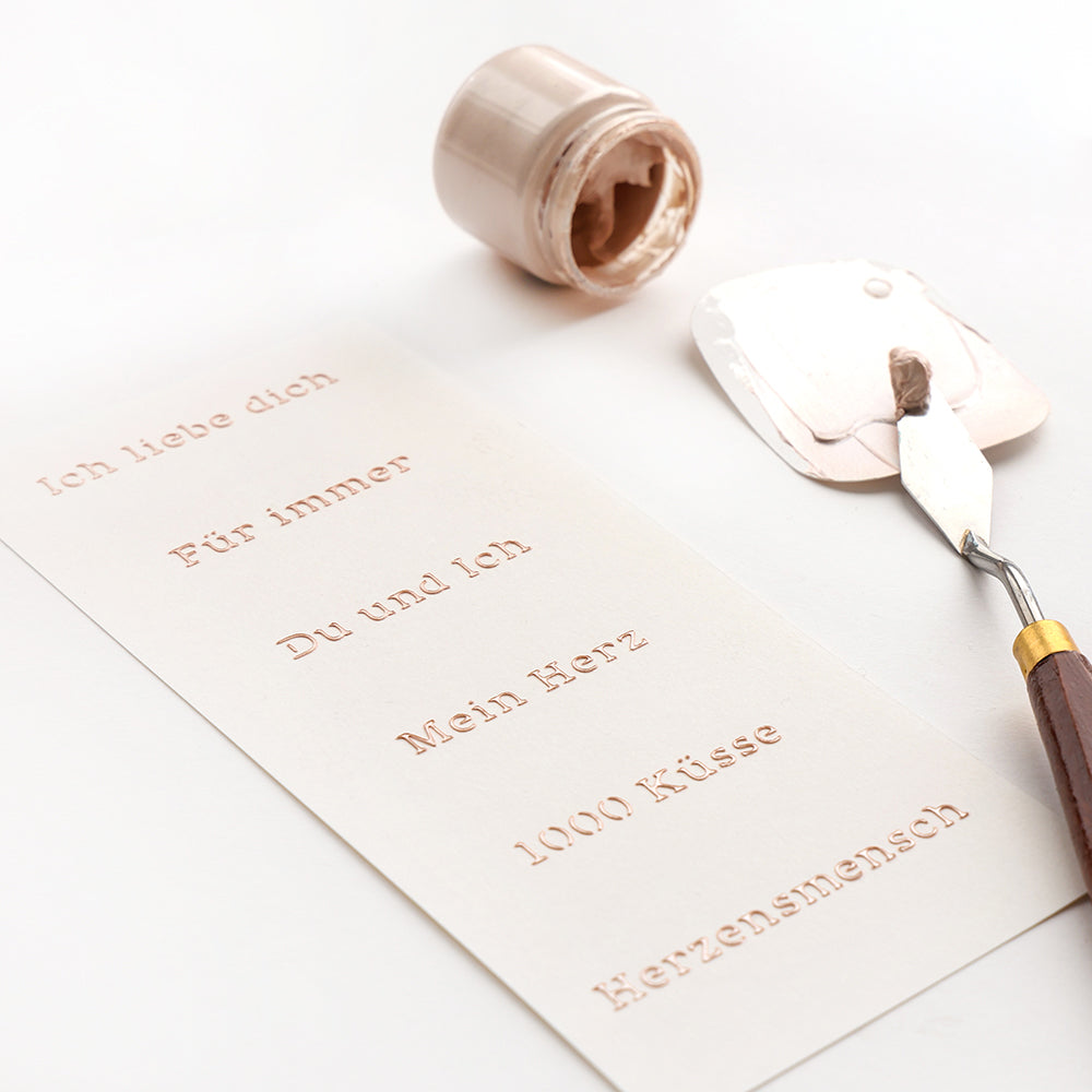 Ein Spachtel mit beiger Farbe liegt neben einer weißen Karte, die mit deutschen Sätzen geprägt ist - perfekt für Alexandra Renke Schablonen Papierprojekte mit wiederverwendbaren Schablonen wie der Schablone Du und Ich.