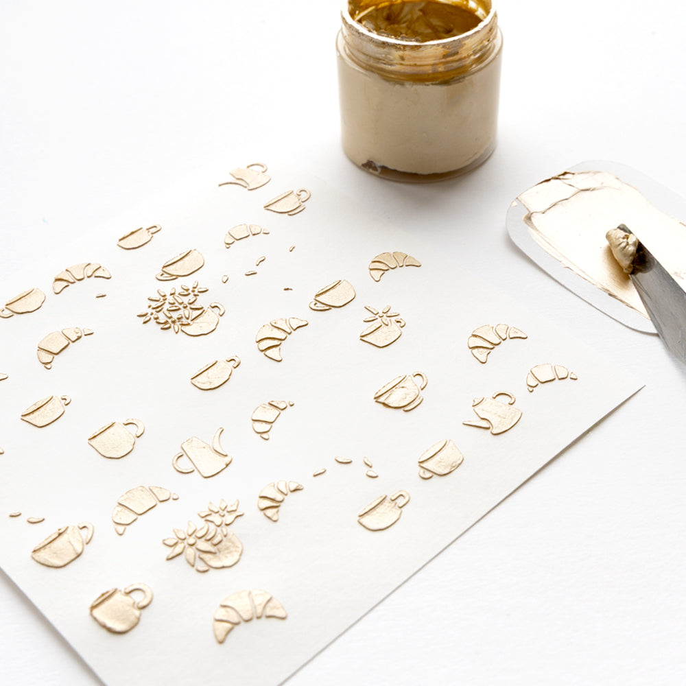 Goldgeprägte Muster von Tassen, Croissants und Blumen auf weißem Papier, hergestellt mit der Alexandra Renke wiederverwendbaren Schablone Florale Kaffeezeit und Goldpaste mit einem Spachtel.