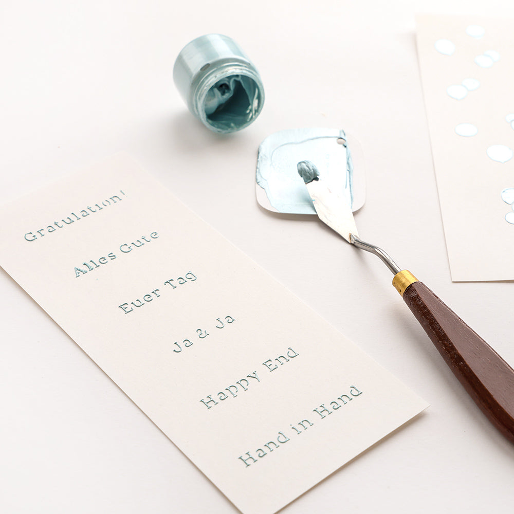 Ein Spachtel mit blauer Farbe neben Karten mit deutschen und englischen Grüßen, die mit einer wiederverwendbaren Alexandra-Renke-Schablone für einfache Bastelarbeiten hergestellt wurden.