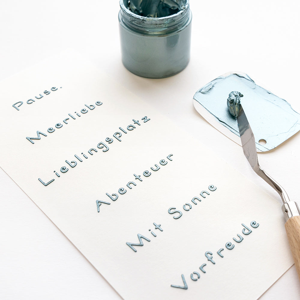 Ein Spachtel und ein Farbtopf neben einem Papier mit deutschen Wörtern in blauer Farbe, die mit wiederverwendbaren Alexandra Renke-Schablonen wie der Schablone Meerliebe erstellt wurden.