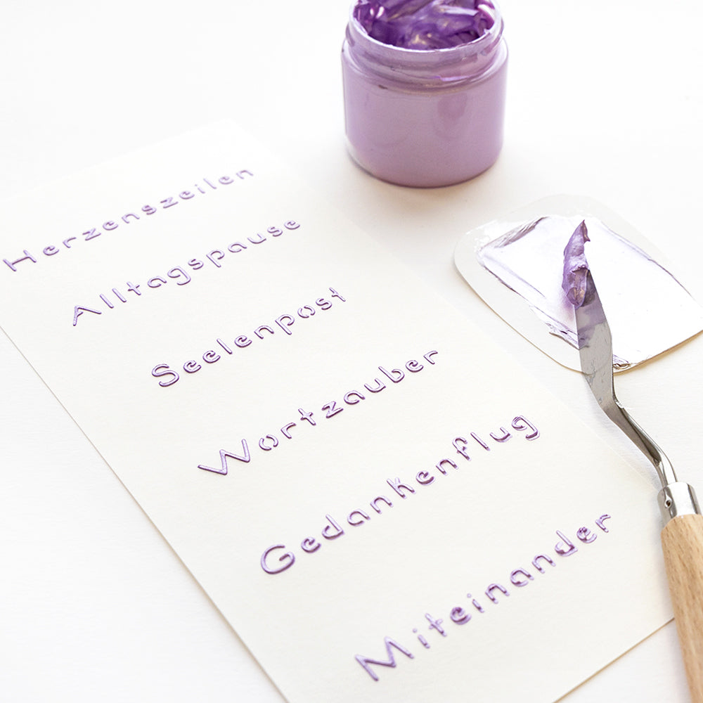 Ein Spachtel mit violetter Farbe ruht neben deutschen Wörtern in erhabenen violetten Buchstaben, die mit Alexandra Renke wiederverwendbaren Schablonen für einzigartige Stencil Wortzauber Effekte auf Papier hergestellt wurden.