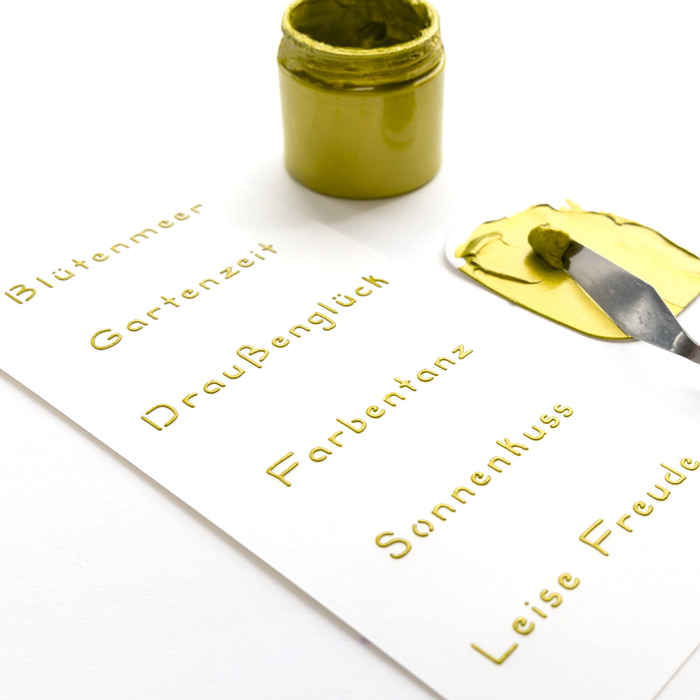 Ein Spachtel und Goldfarbe von Alexandra Renke verleihen einem Papierbogen, der mit wiederverwendbaren Schablonen in Gold geprägte deutsche Wörter enthält, Glanz.