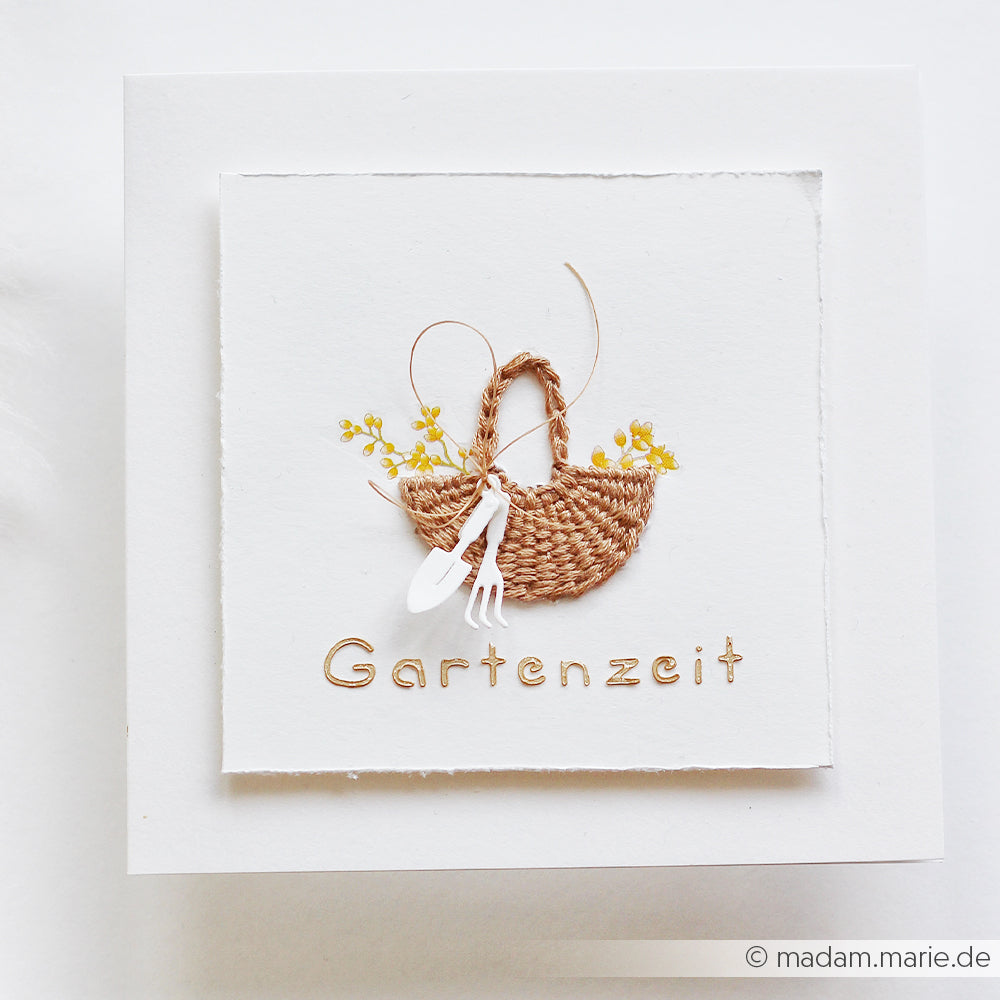 Ein geflochtener Korb mit gelben Blumen und Gartengeräten über der Aufschrift „Gartenzeit“ auf weißem Papier, von Alexandra Renke mit wiederverwendbaren Schablonen gestaltet, was einen bezaubernden Effekt erzeugt.