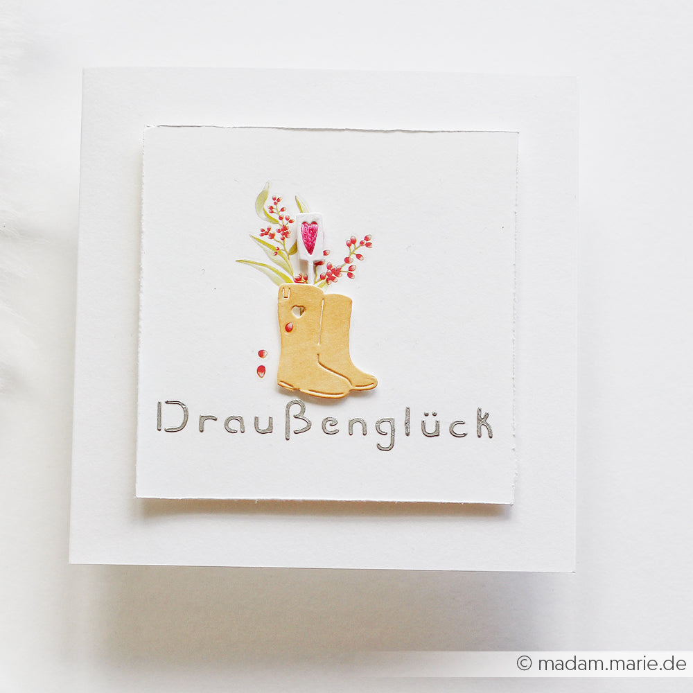 Handgefertigte Karte von Alexandra Renke mit gelben Stiefeln, Blumen und der Aufschrift „Draußenglück“ auf weißem Hintergrund – ideal für Schablonen-Papierprojekte oder mit wiederverwendbaren Schablonen während der Gartensaison.