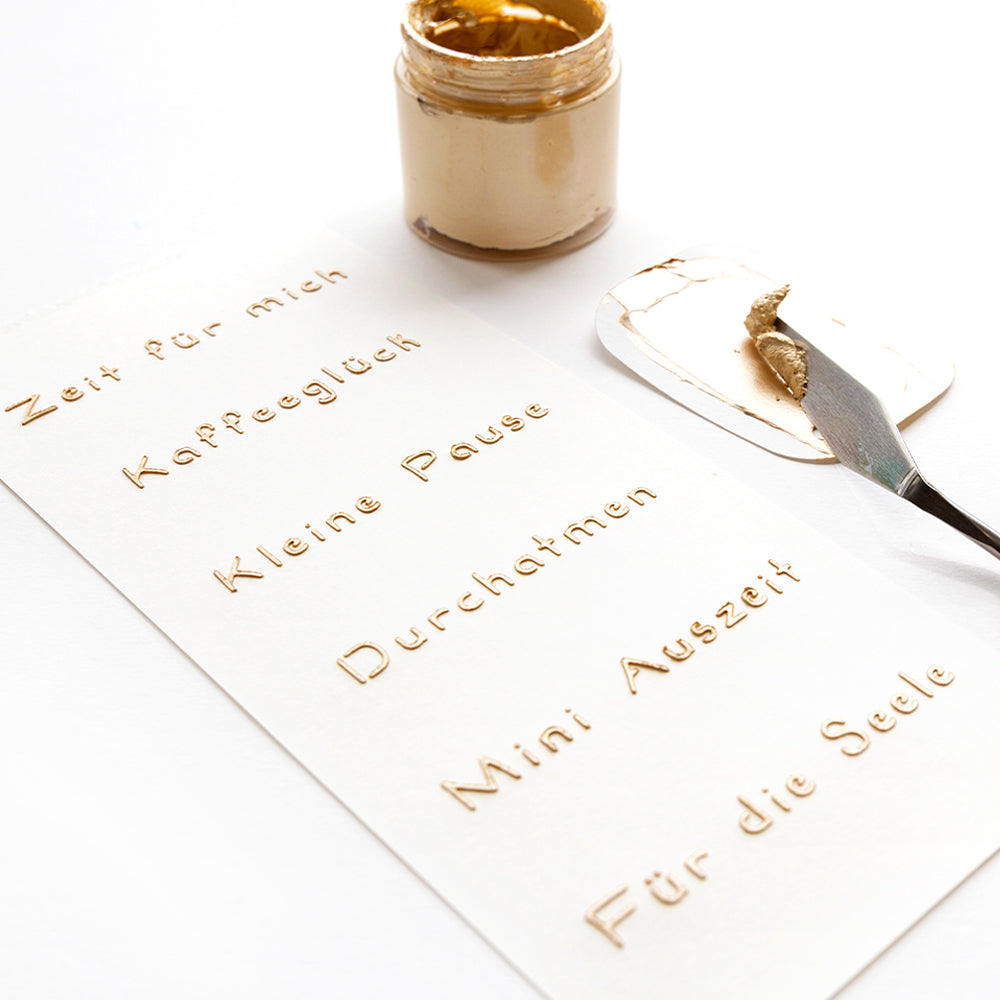 Mit einem Spachtel wird Goldpaste neben deutschen Sprüchen in Gold auf weißem Papier aufgetragen. Alexandra Renke Schablonen Papierprojekte sorgt für kreative, wiederverwendbare Schablonendesigns.