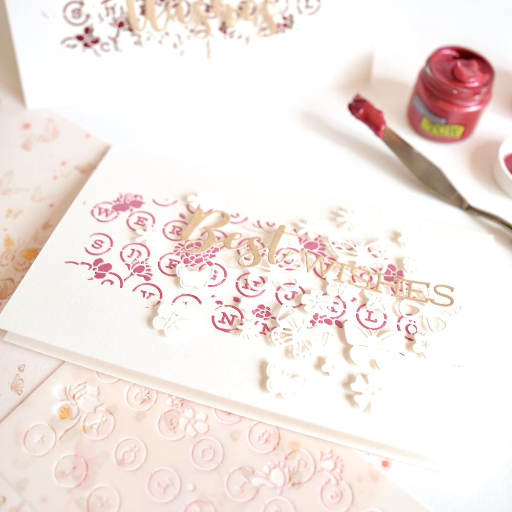 Eine handgefertigte Glückwunschkarte mit rosa Farbakzenten und floralen Ausschnitten, die mit Schablonen von Alexandra Renke und der „Stencil Florale Schreibmaschine“ gestaltet wurden, sowie weiteren hochwertigen Bastelmaterialien, die liebevoll auf einem weißen Untergrund angeordnet sind.