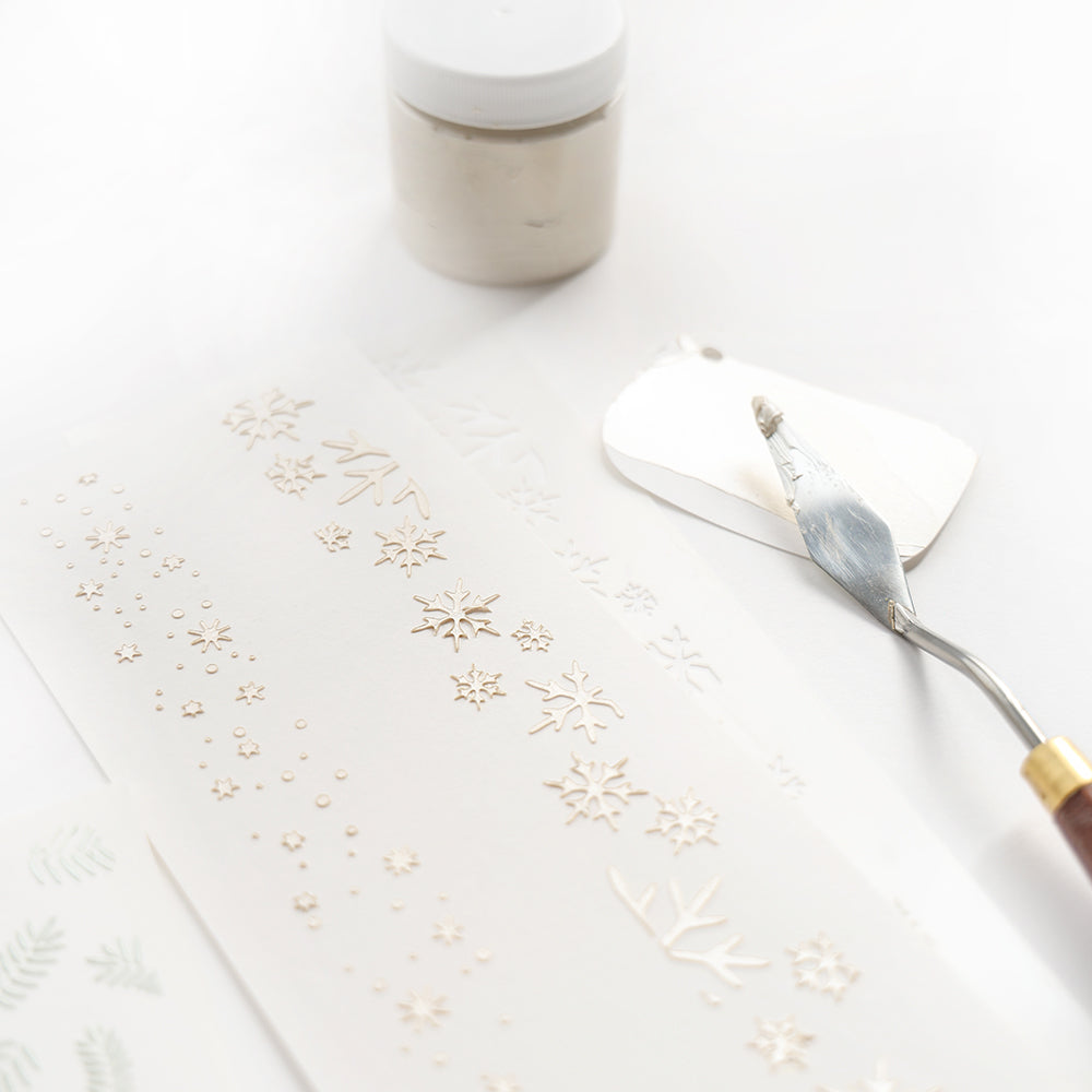 Palettenmesser und Dose neben weißem Papier mit erhabenen Schneeflockenmotiven, hergestellt mit Alexandra Renke wiederverwendbaren Stencils - ideal für Schablonen Papierprojekte und kreative Stencil Schneefall Effekte.