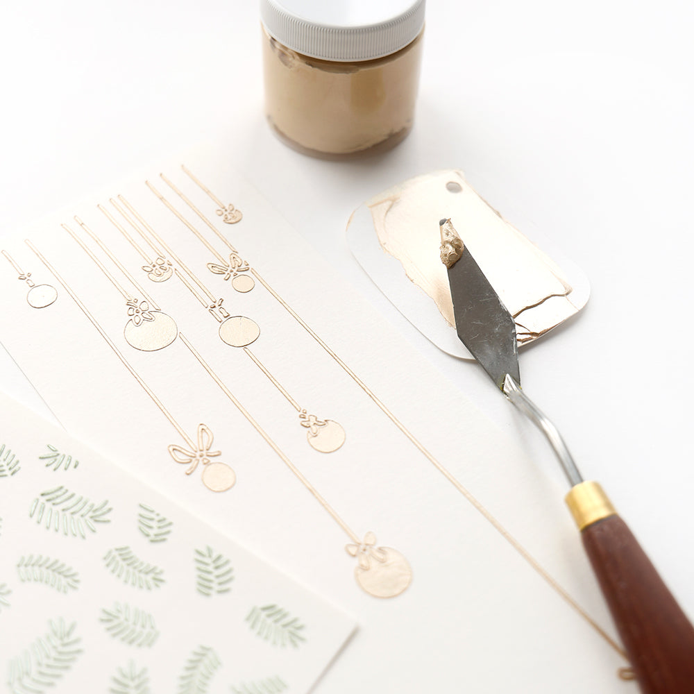 Goldprägepaste und Spachtel neben Prägepapier, einem Bogen mit grünen Blattmotiven und Alexandra Renke wiederverwendbare Schablonen Stencil Festliche Christbaumkugeln für festliche Bastelprojekte.