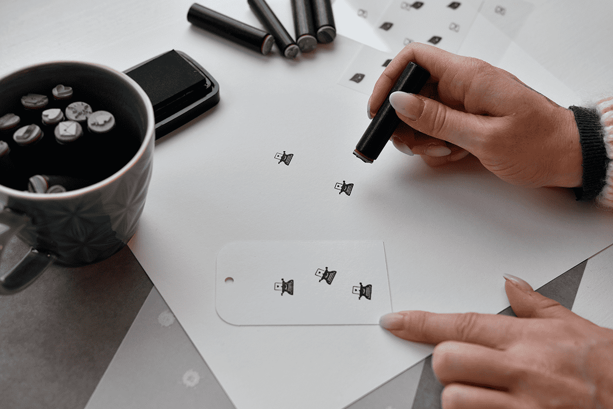 Eine Person stempelt an einem Schreibtisch mit Stempeln, Stempelkissen und einem Becher kleine Hütchensymbole auf Papieranhänger.