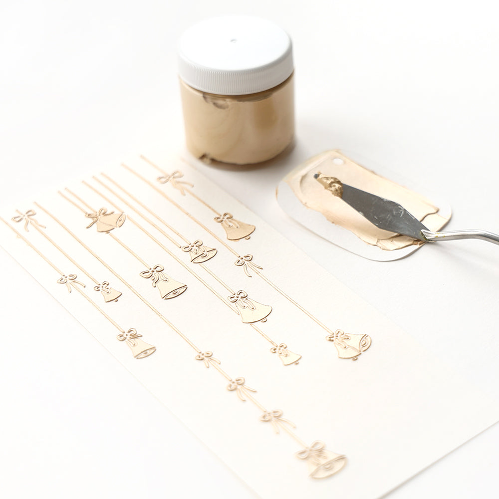 Mit einem Spachtel wird Goldpaste neben einem Papier mit geprägten goldenen Glocken und Schleifen aufgetragen.