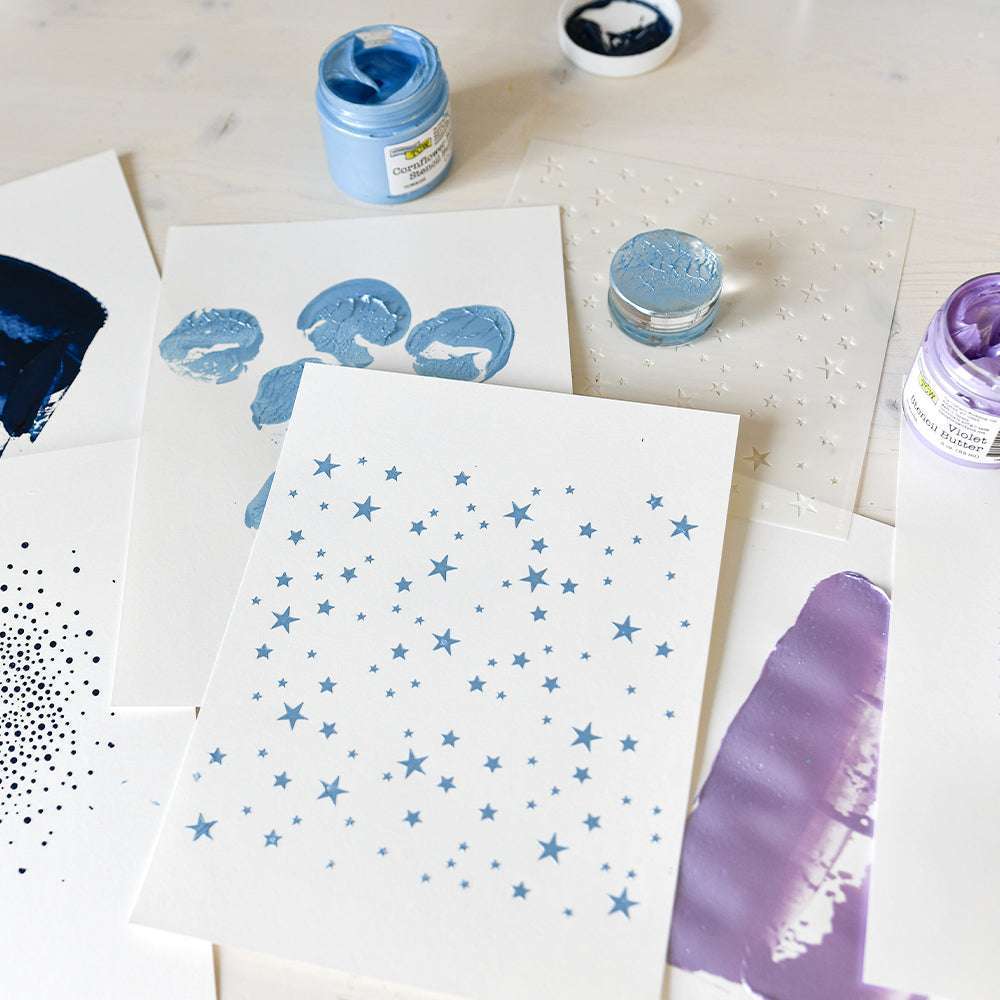 Die Crafters Workshop-Bögen sind mit blauer und violetter Farbe bemalt, darunter einer mit einem blauen Sternmuster, der einen kreativen Mixed-Media-Stil auf einer weißen Oberfläche zeigt.