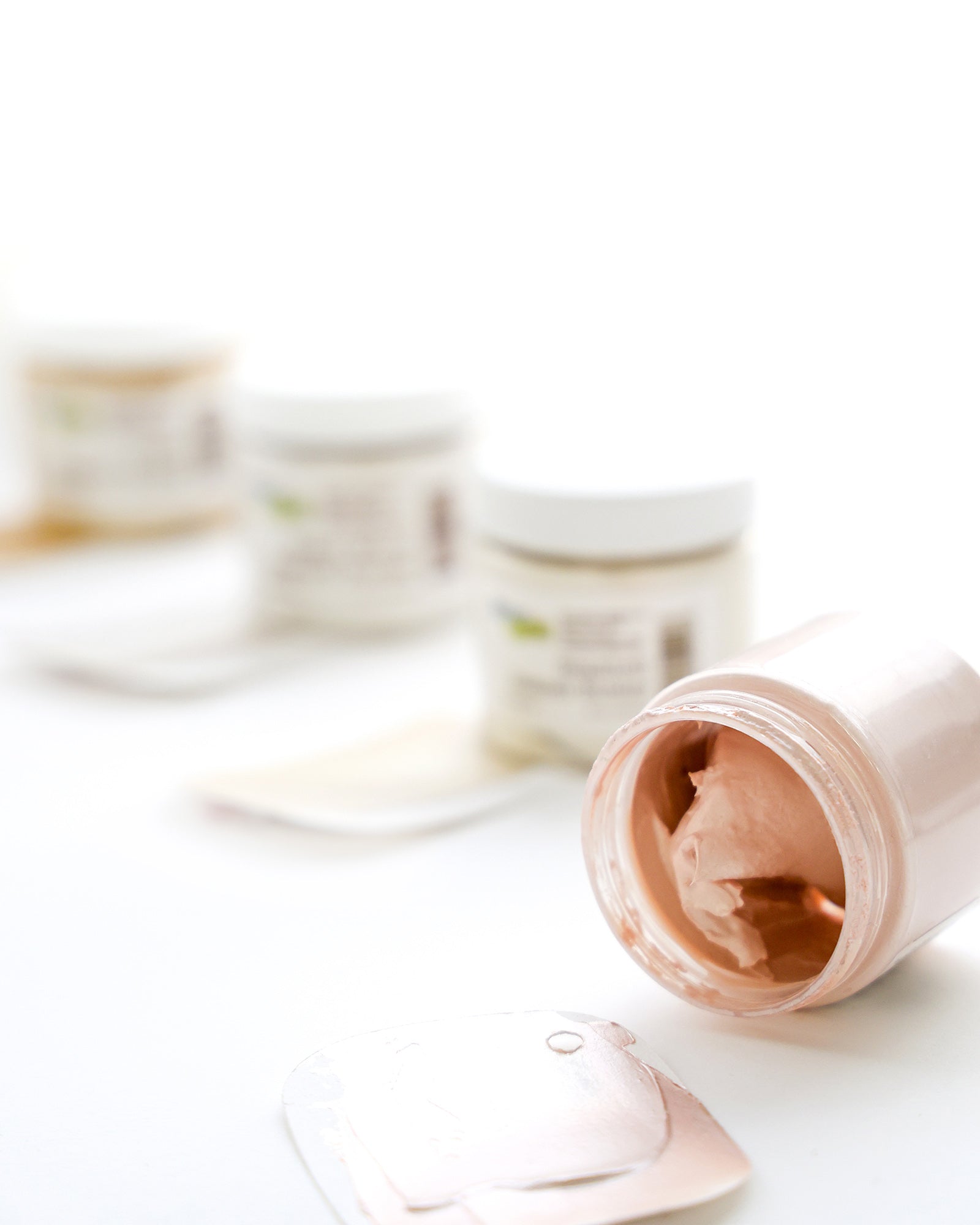 Ein geöffnetes Glas mit rosafarbener Sahne im Fokus, mit drei unscharfen Gläsern im Hintergrund auf einer weißen Fläche.