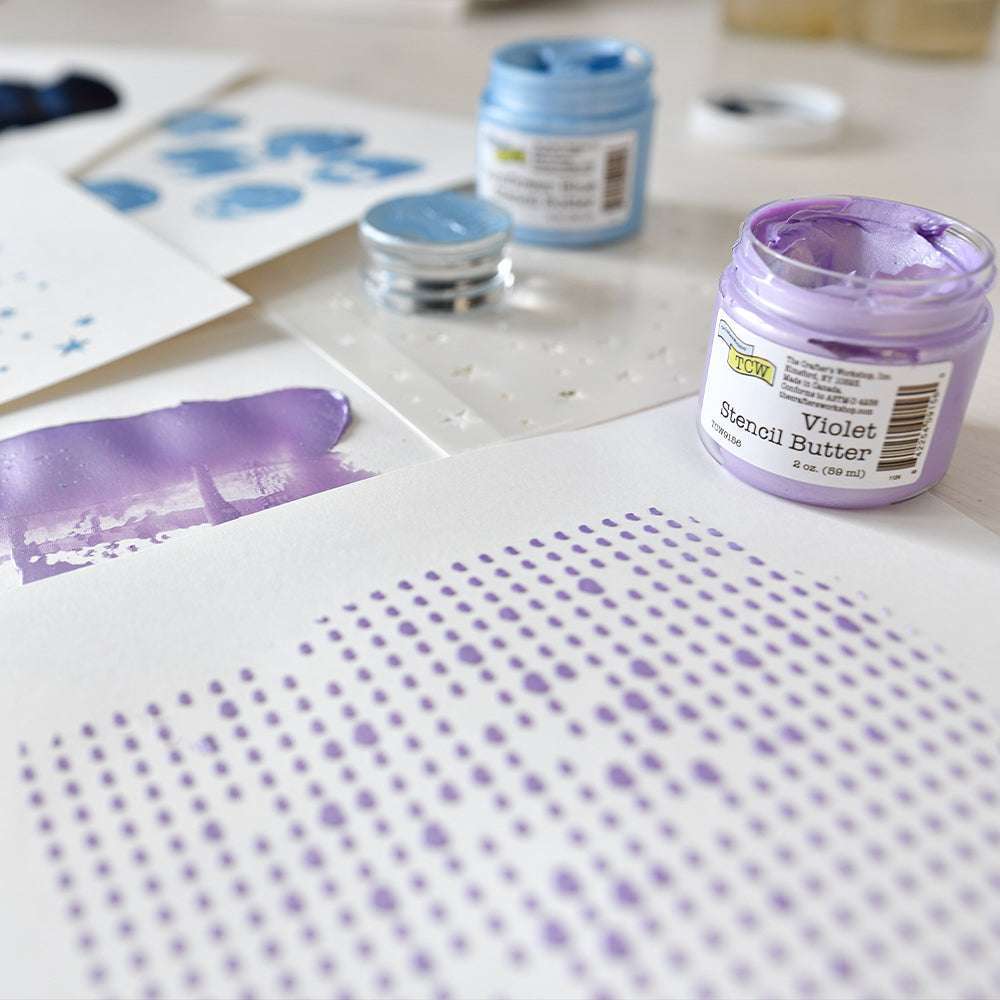 Geöffnete Gläser mit blauer und violetter Stencil Butter von The Crafters Workshop liegen neben gemalten und gepunkteten Schablonen auf weißem Papier, ideal für Mixed Media Projekte.