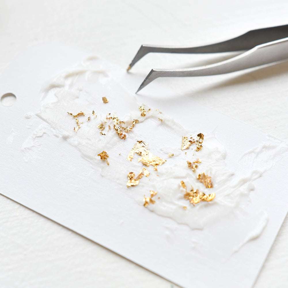 Eine Pinzette mit Blattgoldflocken und Tacky-when-Dry Gel von The Crafters Workshop auf einem weißen Anhänger - ideal, um Mixed-Media-Projekten strukturierte Effekte zu verleihen.