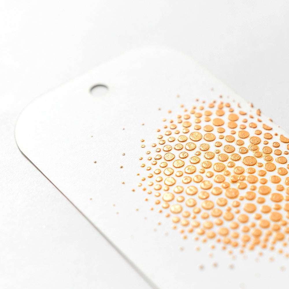 Ein weißer Anhänger von The Crafters Workshop weist eine Ansammlung erhabener, glänzender goldener Punkte auf, die mit Schablonenbutter oder Modellierpaste erzeugt wurden, sowie ein Loch am oberen Rand.