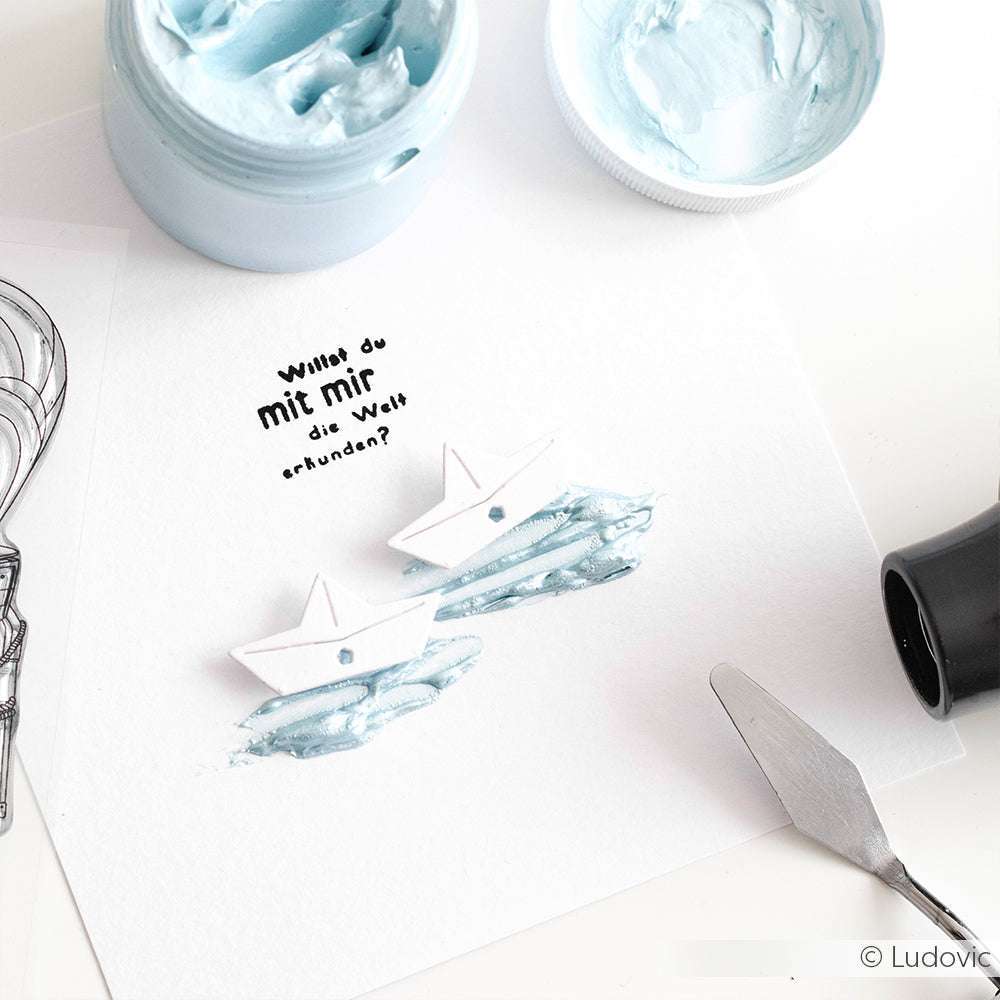 Zwei Papierboote auf strukturierter blauer Farbe, erstellt mit Mischtechniken unter Verwendung von The Crafters Workshop Kunstwerkzeugen, Modellierpaste, deutschem Text und einem Glas hellblauer Farbe in der Nähe.