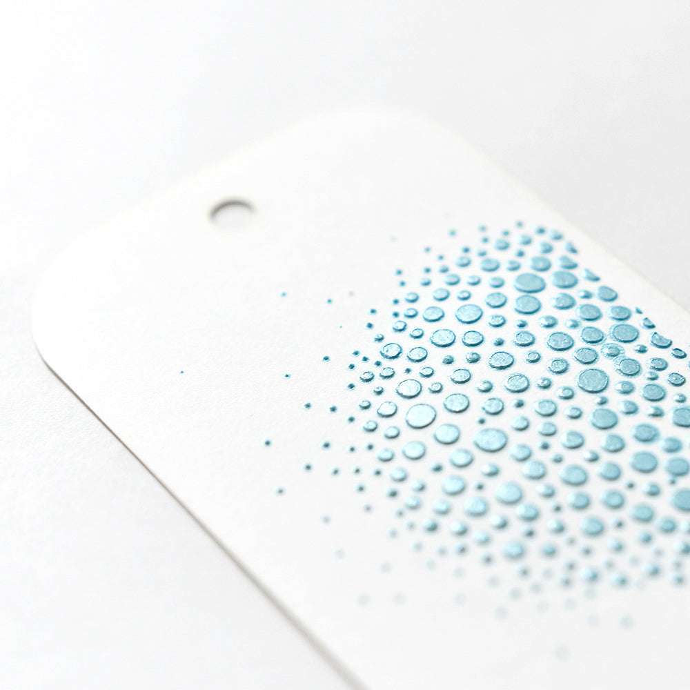 Ein weißer Anhänger von The Crafters Workshop mit einem Loch und erhabenen blauen Punkten in einem Farbverlauf - ideal, um Stencil Butter hinzuzufügen oder andere Mischtechniken in Ihren kreativen Projekten anzuwenden.