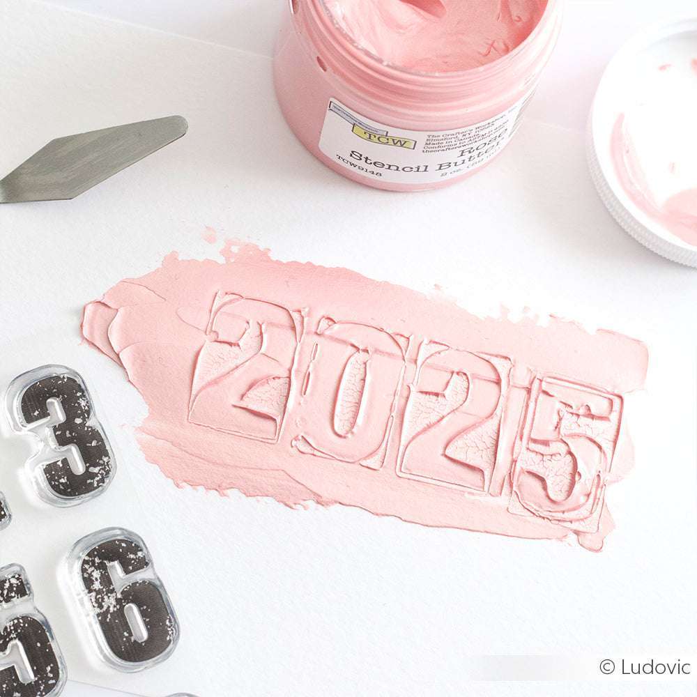 Die rosafarbene Schablonenbutter von Crafters Workshop buchstabiert "2025" auf weißem Papier, mit Schablonennummern und einem Spachtel - ideal zum Hinzufügen von Texturen zu Mixed-Media-Tafeln oder zur Kombination mit Modellierpaste für einzigartige Effekte.