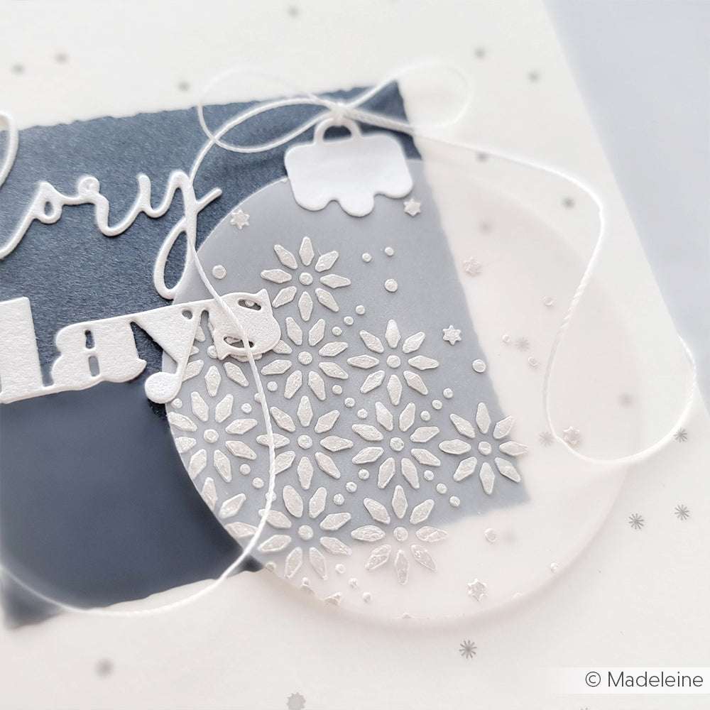 Nahaufnahme einer handgefertigten Ornamentkarte mit weißen geprägten Schneeflocken, einer Schleife und grauem Hintergrund - ideal für Mixed-Media-Projekte mit The Crafters Workshop Stencil Butter und Schablonen, um Textur hinzuzufügen.