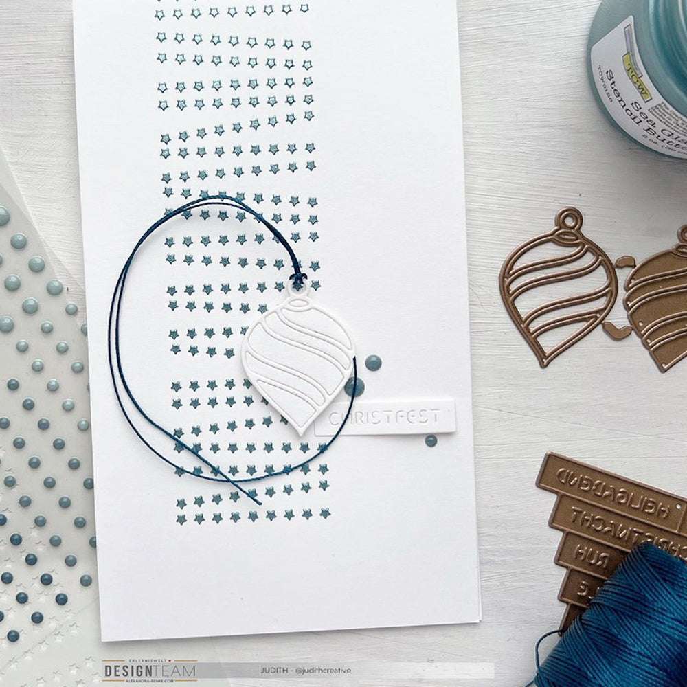 Handgefertigte Weihnachtskarte mit blauen Punkten, einem aufgereihten Ornament und Bastelmaterial auf weißem Untergrund. Die Mixed-Media-Techniken von The Crafters Workshop mit Modellierpaste sorgen für zusätzliche Textur und kreatives Flair.
