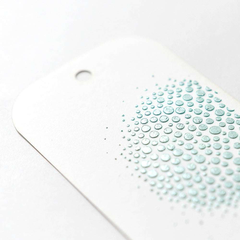 Ein weißes Etikett mit abgerundeten Ecken weist einen Farbverlauf aus tealfarbenen Punkten auf, der mit Stencil Butter von The Crafters Workshop für einen strukturierten Mixed-Media-Look erstellt wurde (siehe Nahaufnahme).