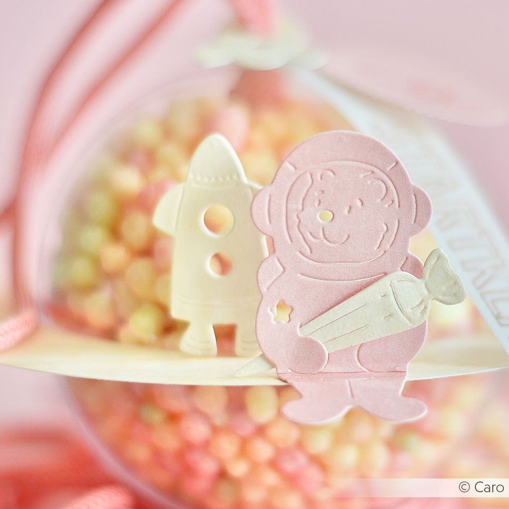 Die rosafarbenen und cremefarbenen Astronauten- und Raketenausschnitte von Alexandra Renke werden mit bunten Zuckerperlen in einem runden, durchsichtigen Behälter dahinter präsentiert.