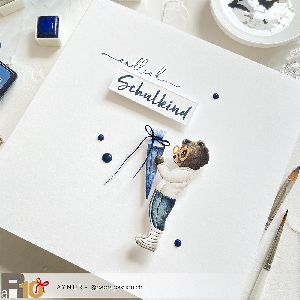 Ein Bär in Jeans hält eine blaue Schultüte neben dem Schriftzug "Endlich Schulkind" auf einer weißen Karte, die mit den Stanzwerkzeugen Motivstanze Bär mit Schultüte und Stanzen für Designpapier von Alexandra Renke hergestellt wurde.