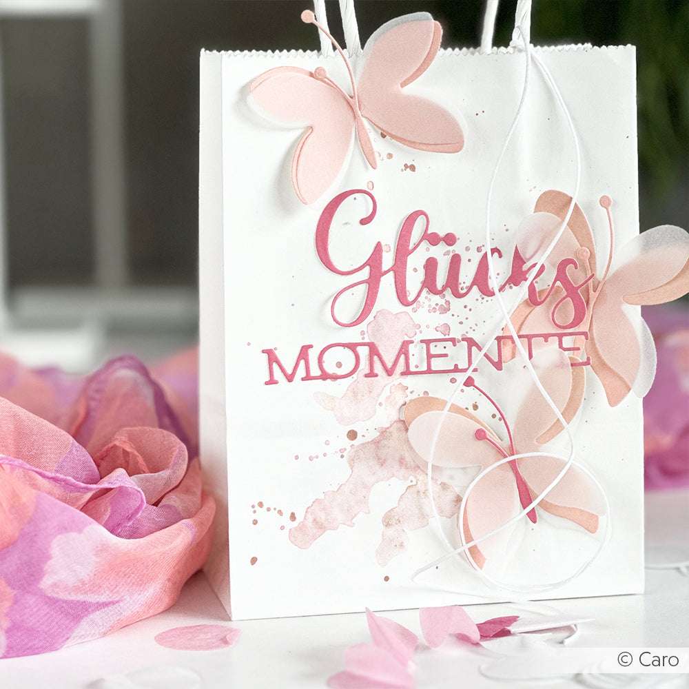 Eine weiße Geschenktüte mit rosafarbenen Schmetterlingen, die mit Alexandra Renke Motivstanze Schichtschmetterling und "Glücks Momente" in rosafarbener Schrift ausgestanzt wurde.