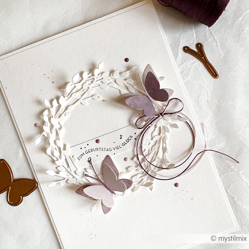 Handgefertigte Geburtstagskarte von Alexandra Renke mit weißem Kranz, lilafarbenen Schmetterlingen, die mit Motivstanze Schichtschmetterling DieCut Systeme hergestellt wurden, einer Schleife und deutschen Geburtstagswünschen.