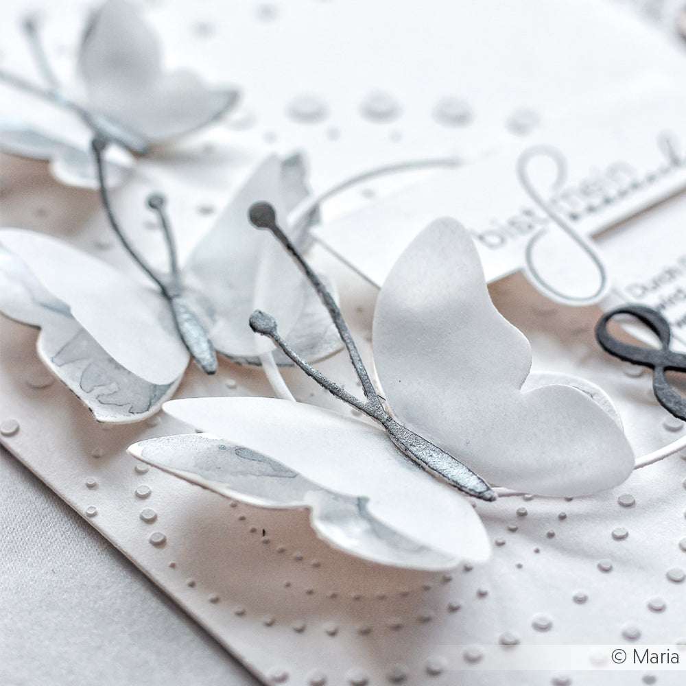 Nahaufnahme von weißen Papierschmetterlingen, hergestellt mit Alexandra Renke Motivstanze Schichtschmetterling, dargestellt auf einer strukturierten, gepunkteten weißen Karte mit eleganten Hintergrunddetails.