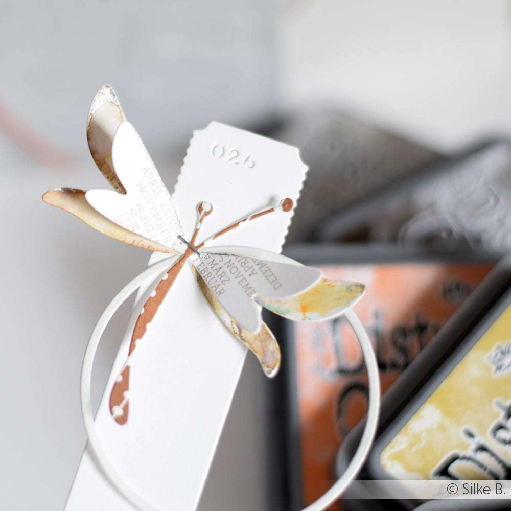 Ein weißes Lesezeichen von Alexandra Renke ist mit einer Metallic-Libelle aus der Motivstanze Schichtlibelle verziert, die neben bunten Stempelkissen zu sehen ist.