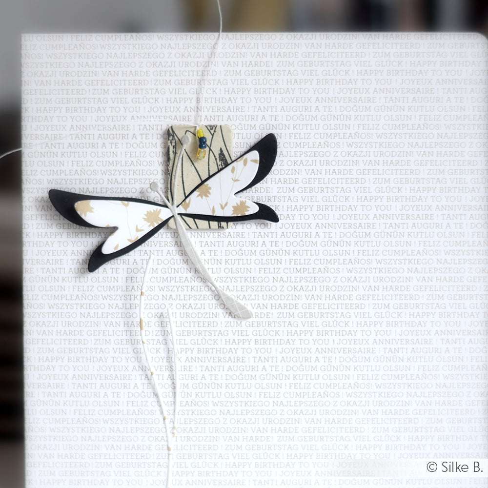Eine mit Alexandra Renke Designpapieren und einer Motivstanze Schichtlibelle gebastelte Papierlibelle hängt an einer Schnur an einer Karte, auf der "Happy Birthday" in mehreren Sprachen steht.