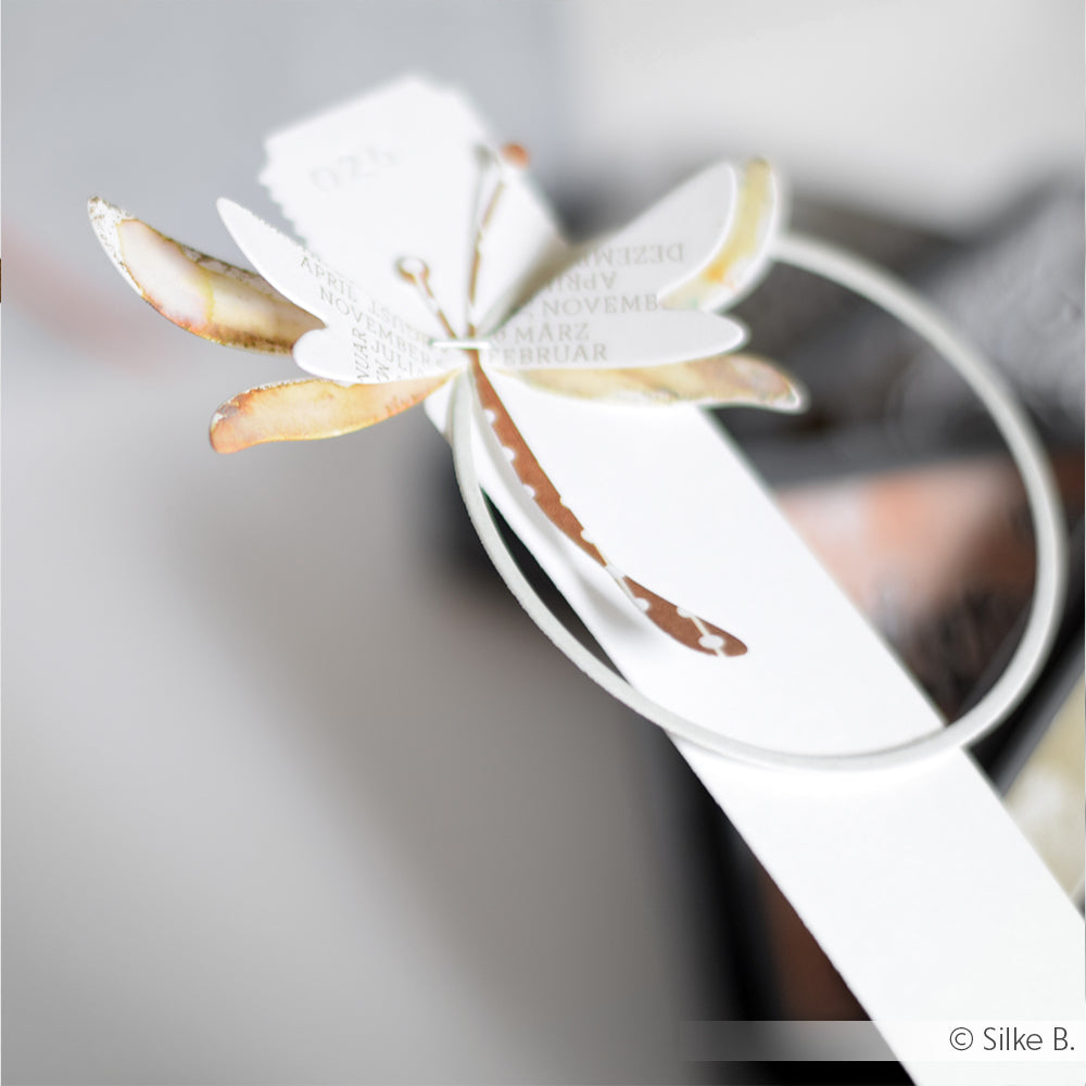 Das mit DieCut Systemen und Designpapieren hergestellte Alexandra Renke Papierlibellen-Lesezeichen zeigt Text auf den Flügeln und ruht auf einer weißen Oberfläche.