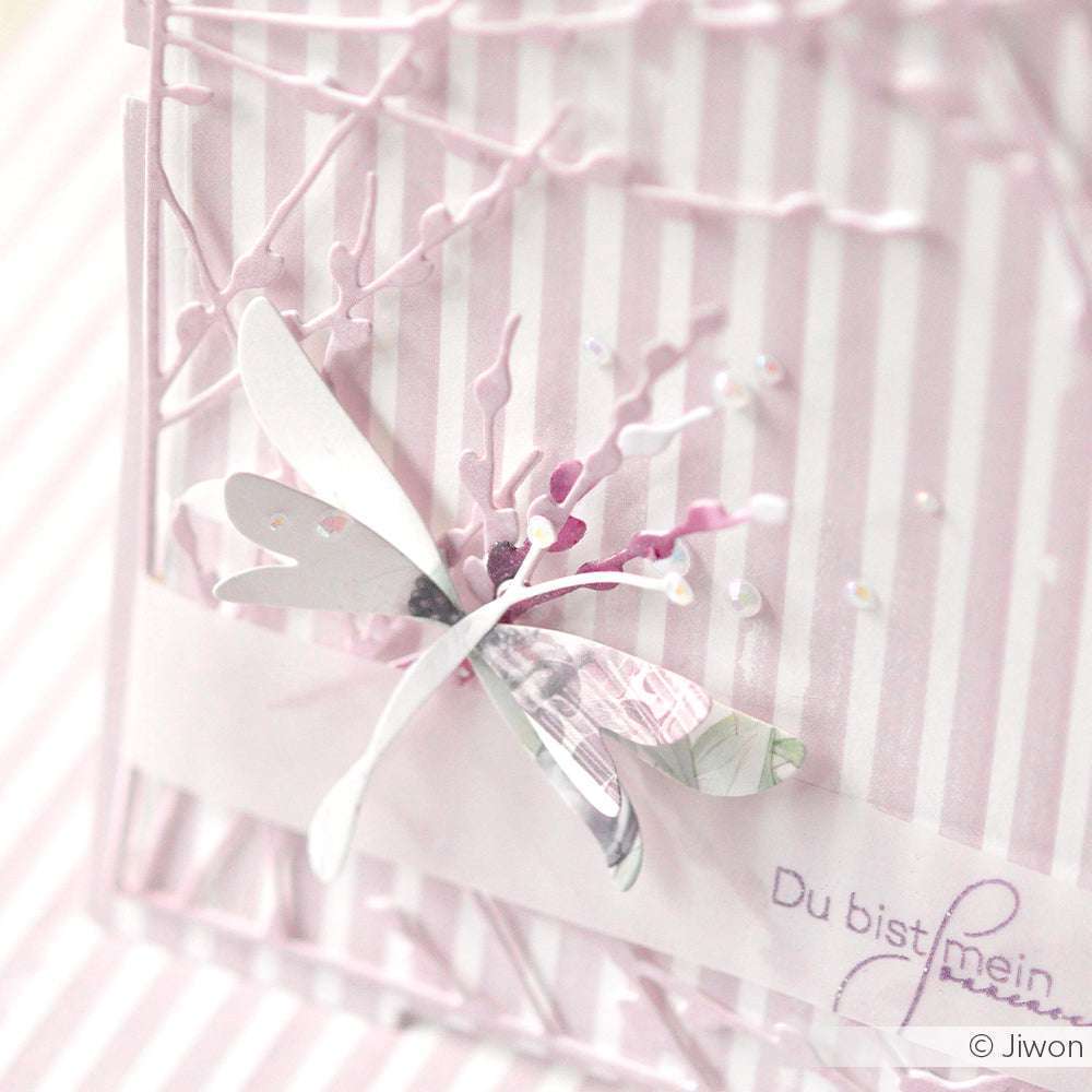 Eine zarte Papierlibelle, hergestellt mit der Alexandra Renke Motivstanze Schichtlibelle Dies, ruht auf einer gestreiften pastellrosa und weißen Karte mit deutschem Text.