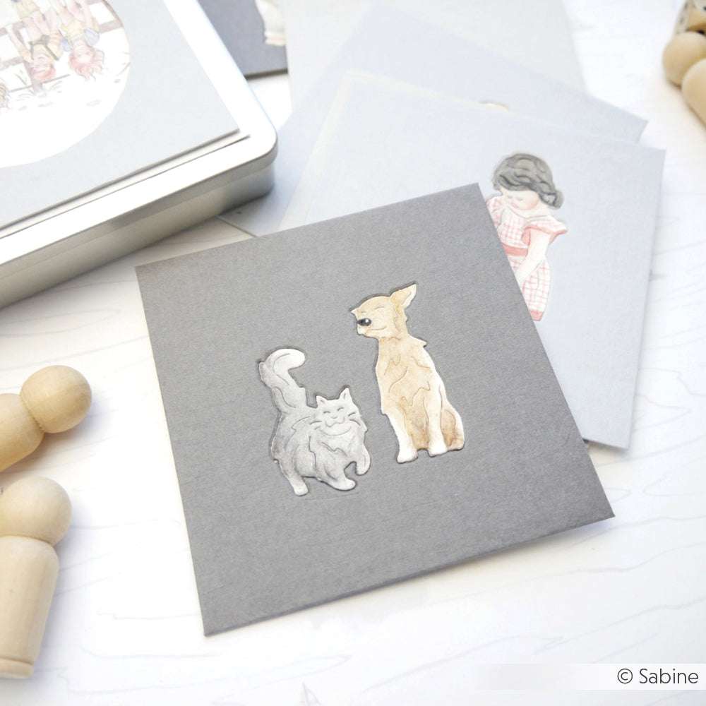 Eine graue Karte von Alexandra Renke mit niedlichen Katzen- und Hundeillustrationen aus der Motivstanze ist von Holzpflöcken und Papiergegenständen umgeben.