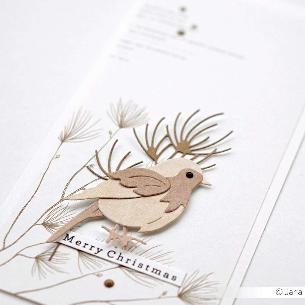 Ein Papiervogel, der mit den Alexandra Renke Motivstanze Rotkehlchen ausgestanzt wurde, sitzt auf illustrierten Tannenzweigen mit dem Text "Frohe Weihnachten" auf einer weißen Karte.