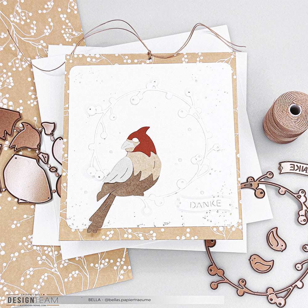 Eine handgefertigte Karte von Alexandra Renke zeigt einen rot-braunen Vogel aus der Motivstanze Schichttier Kardinal, der auf einem weißen Zweig sitzt und mit Designpapieren und Bastelmaterial verziert ist.