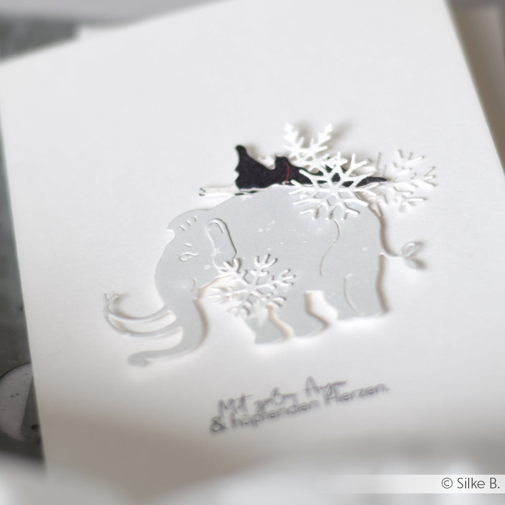 Eine weiße Karte mit den Motivstanzen Mammut Familie von Alexandra Renke zeigt einen gestanzten Elefanten, verziert mit Schneeflocken und einem kleinen schwarzen Vogel, der auf dem Elefanten sitzt.