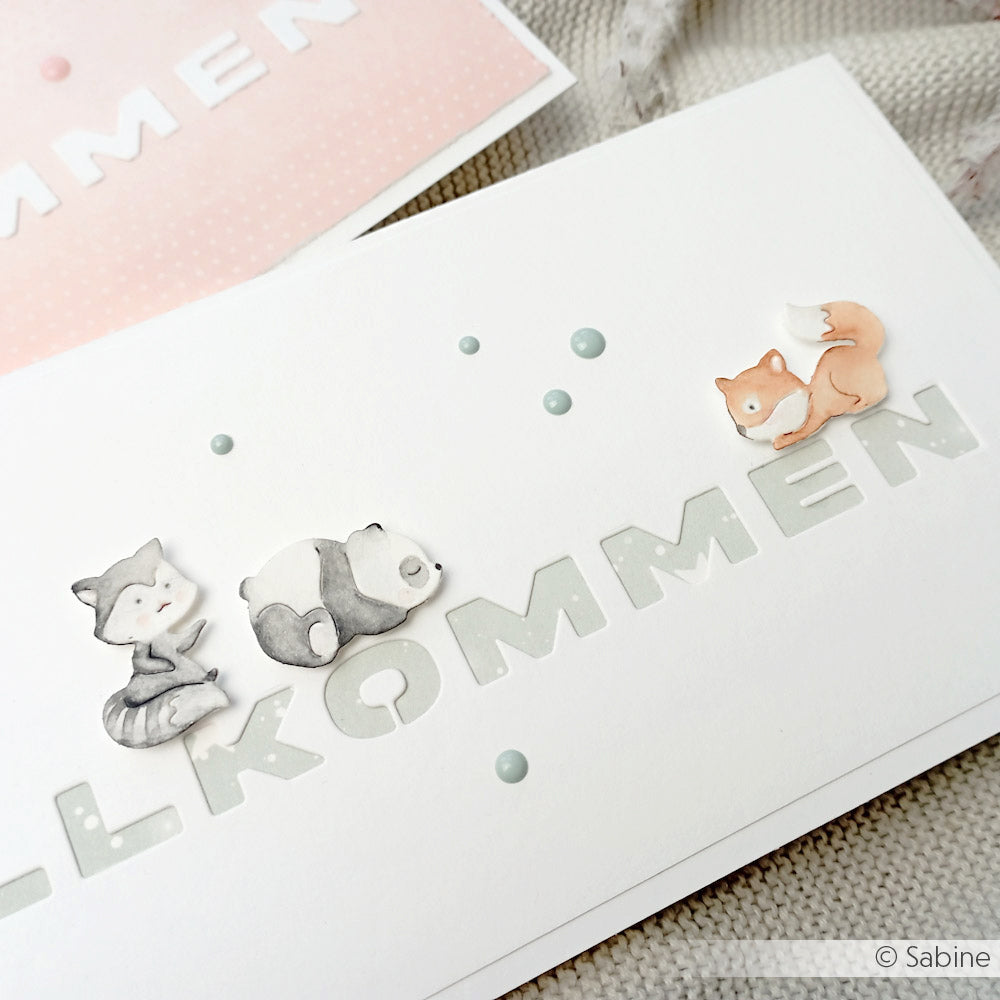 Weiße Karte von Alexandra Renke mit pastellfarbenem WILLKOMMEN-Text und niedlichen Waschbär-, Panda- und Fuchs-Illustrationen aus dem Motivstanze Mini Tier Set Stanzsysteme.