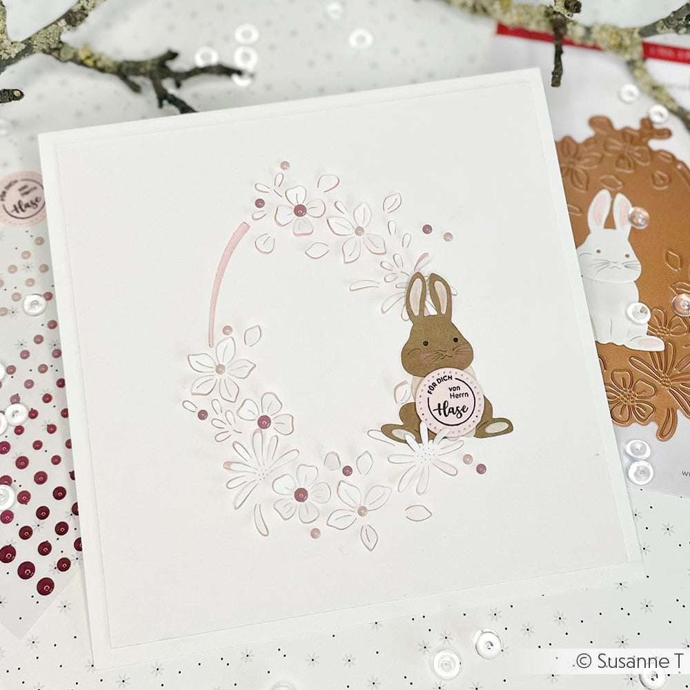 Handgemachte Karte von Alexandra Renke mit einem Hasen, der mit Motivstanze Hase ausgestanzt wurde und ein "Frohe Ostern"-Schild in der Hand hält, umgeben von einem rosa und weißen Blumenkranz.