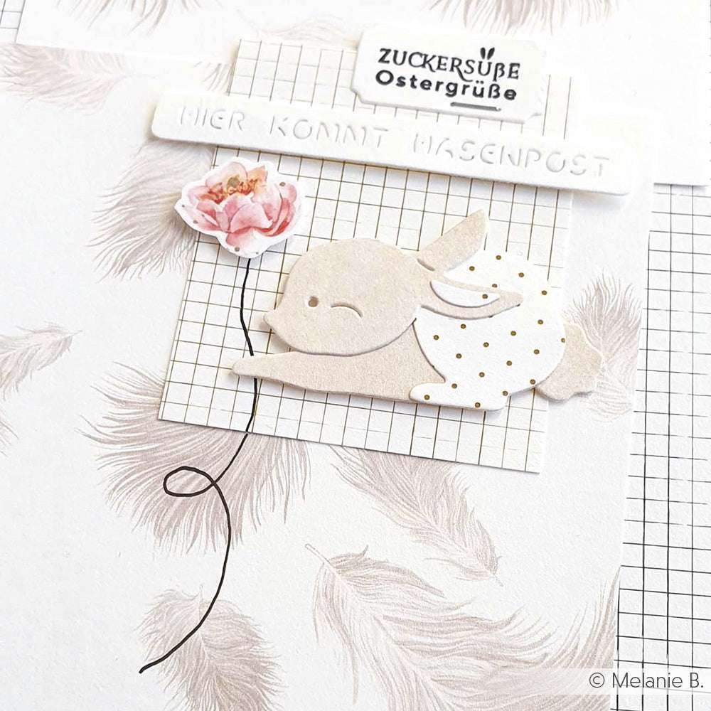 Ein mit der Motivstanze Schichttier Hase von Alexandra Renke gestalteter Papierhase sitzt zwischen Blumen und deutschem Text vor einem hellen Hintergrund mit Federmustern, Gitternetzlinien und eleganten Akzenten von Designpapieren.