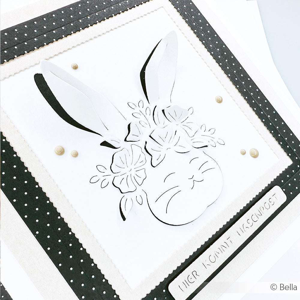 Grußkarte von Alexandra Renke mit einem weißen Hasengesicht und einer Blumenkrone, die mit der Motivstanze Hase ausgestanzt wurde. Die Karte ist mit deutschem Text und einer stilvollen schwarz-weißen Tupfenbordüre verziert, die für zusätzlichen Charme sorgt.