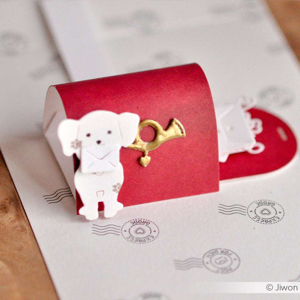 Ein roter Papierbriefkasten mit einem goldenen Horn und einem weißen Hund, der einen Briefumschlag hält, hergestellt mit Alexandra Renke Designpapieren und einer Stanze, dargestellt auf einem Hintergrund mit Buchstabenmuster.