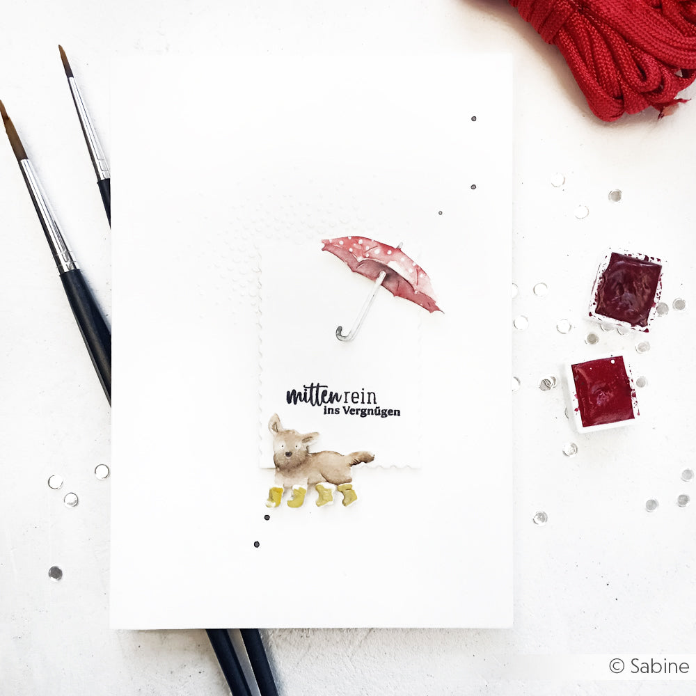 Eine handgefertigte Karte von Alexandra Renke mit einem aquarellierten Hund in gelben Stiefeln mit einem rosa Regenschirm und "mitten rein ins Vergnügen", hergestellt mit der Hundewetter-Stanze und Designpapier-Stanzformen.