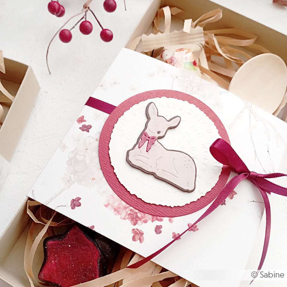 Geschenkpackung mit einem weißen Hirschkeks auf einer Blumenkarte aus Alexandra Renke Designpapier, gebunden mit einem burgunderroten Band und umgeben von festlichem Dekor.