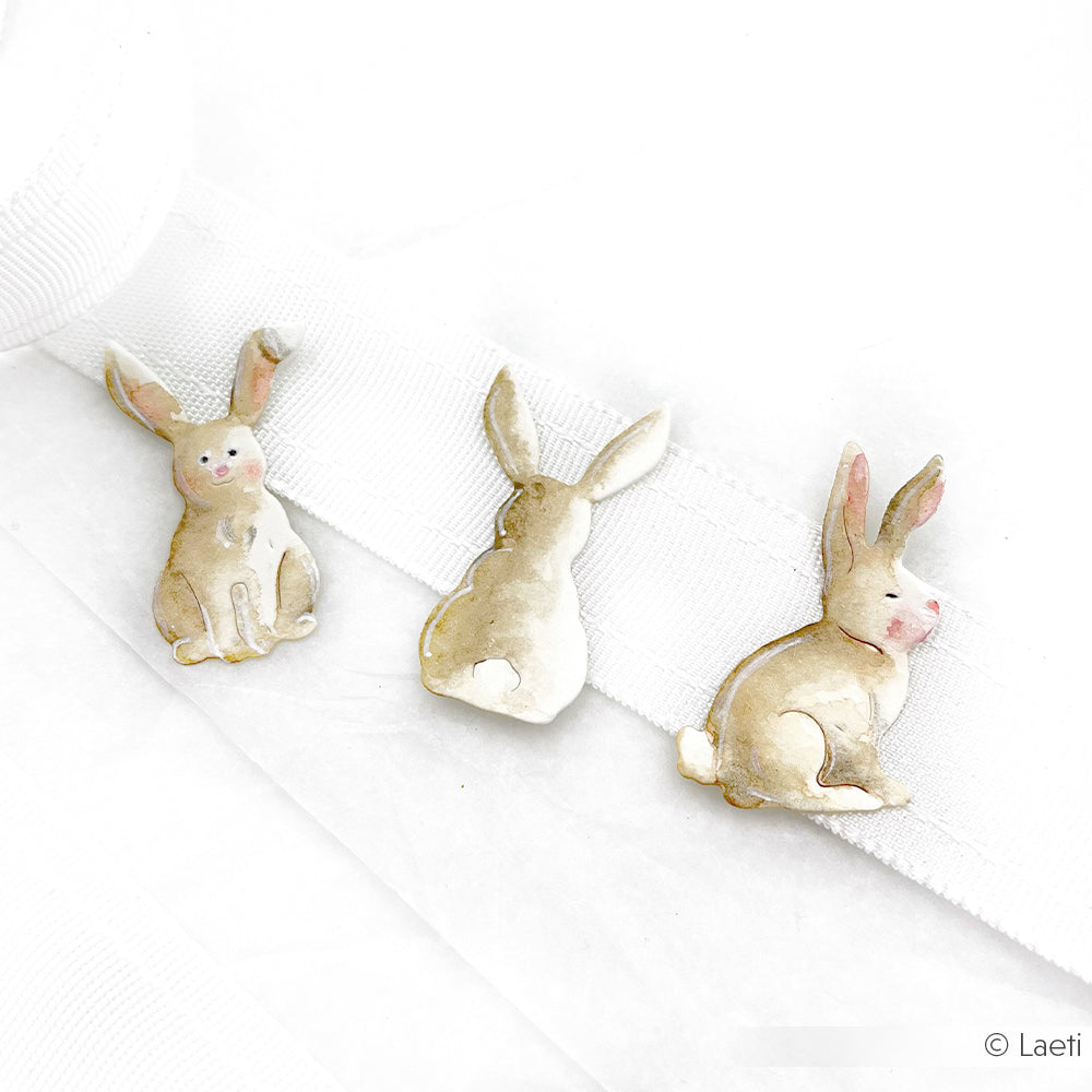 Drei kleine, illustrierte Hasenpins aus der Alexandra Renke PAPPcut®ies Motivstanze Hoppel Trio Kollektion sind in Pastellfarben gehalten und auf einem weißen Band und Hintergrund wunderschön arrangiert.
