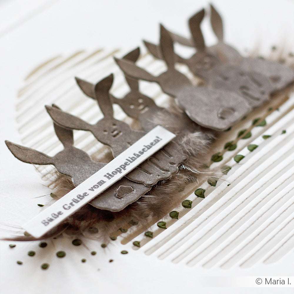 Fünf Papierhasen, die mit der Alexandra Renke Motivstanze Hoppel Trio ausgeschnitten wurden, und ein Streifen mit deutschem Text liegen auf einem strukturierten weißen Hintergrund mit grünen Akzenten.